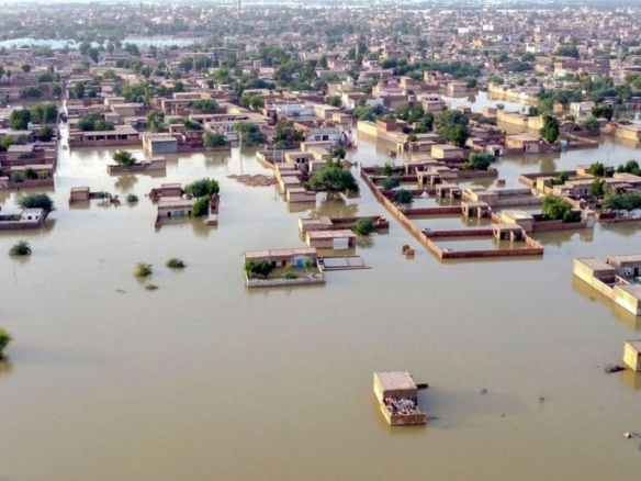 A flooded street in Lahore showing cars submerged in water next to modern housing society buildings.
