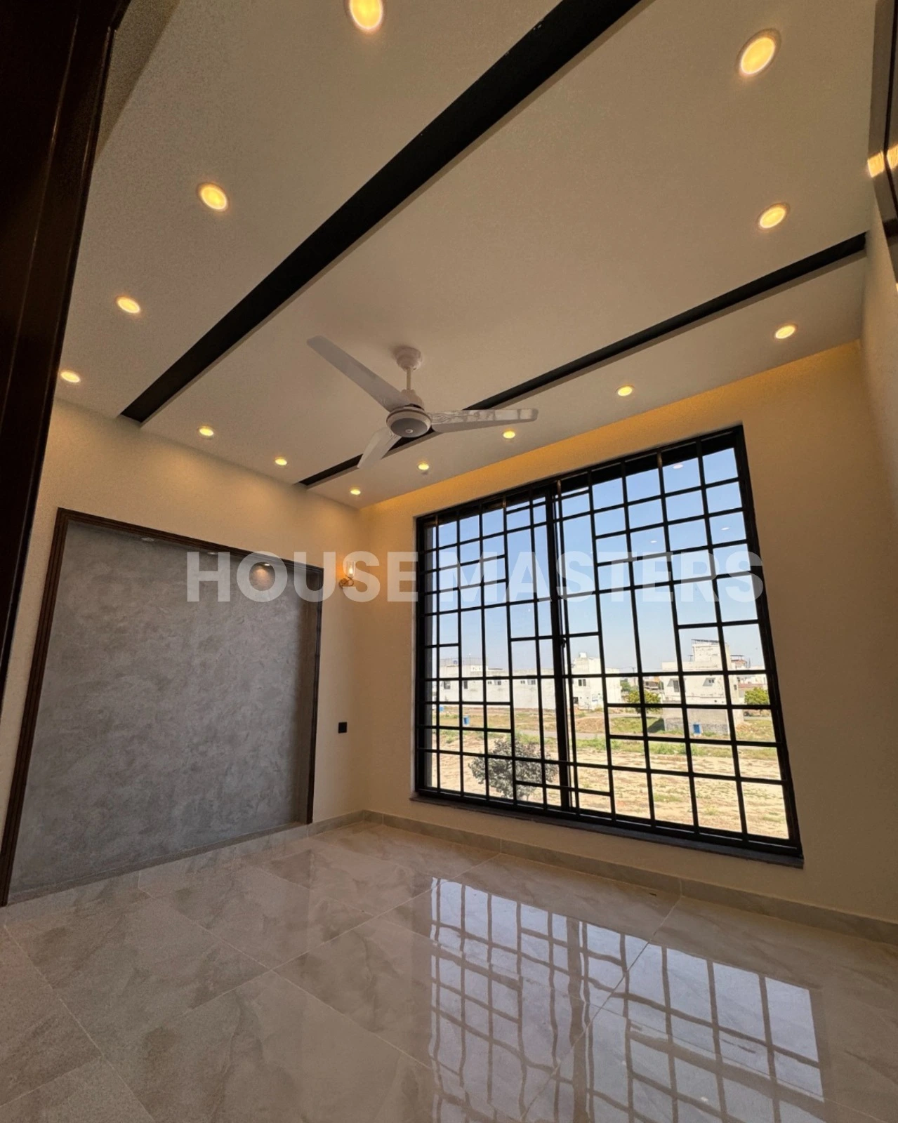 Modern bedroom with large black-framed windows, ceiling fan, and luxurious flooring in a 5 Marla house, offering scenic views in Bahria Orchard, Lahore.
